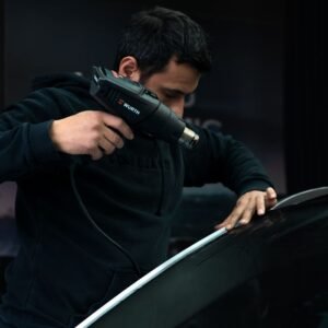 Mechanic carefully applies heat to car windshield with a heat gun in a garage setting.