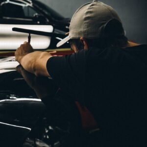 A mechanic wearing a cap examines a car under low light in a garage.